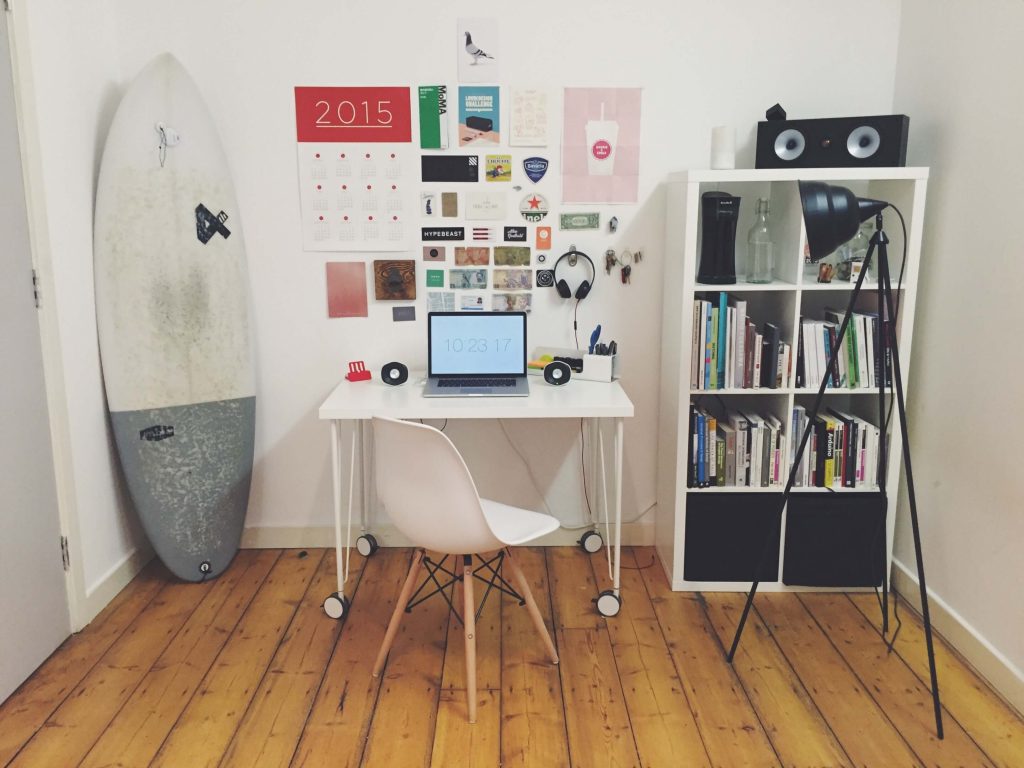 A room with a surfboard, desk and book case.